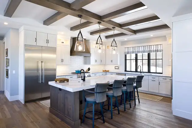 Kitchen remodel with dark wood ceiling beams, glass pendant lights over a white island on a stained wood base, and blue counter stools