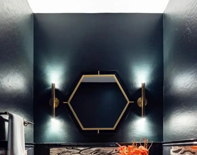 Powder room with a gold hexagon mirror between two brass sconces on a dark painted wall